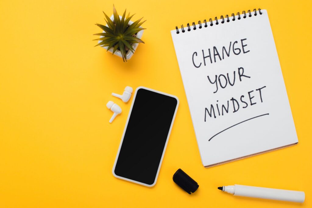 Notebook with change your mindset inscription near smartphone, wireless headphones, potted flower